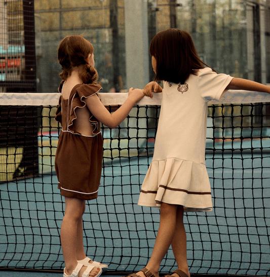 Two sisters in coordinated outfits, one in a brown ruffle romper and one in an ivory pleated polo dress, at a tennis net